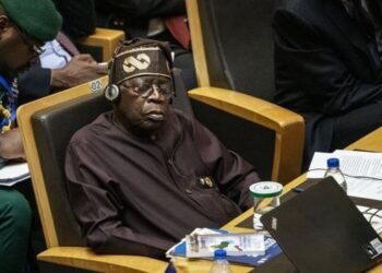 A member of a national delegation seated with eyes closed during a summit session, a Nigerian flag on the table.