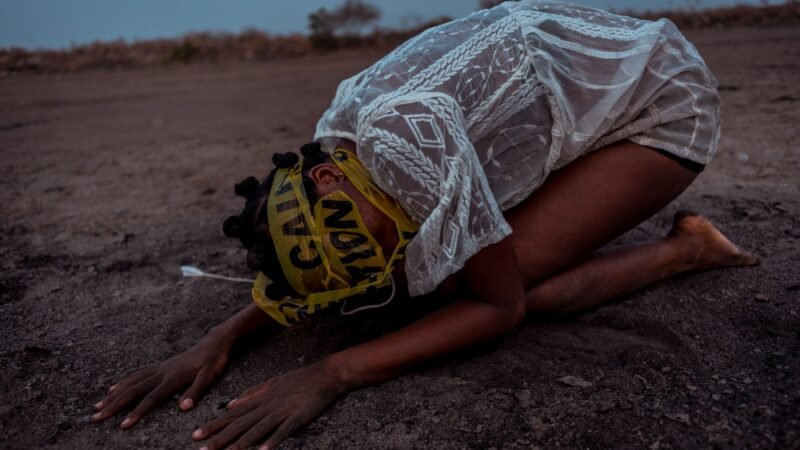 A young climate activist performing a protest piece on cracked dry ground, wearing caution tape.
